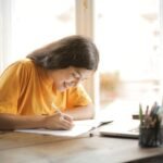 Young woman studying with a laptop and notepad at home, smiling and focused.