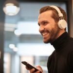 Bearded man smiling with headphones and smartphone indoors, enjoying music.